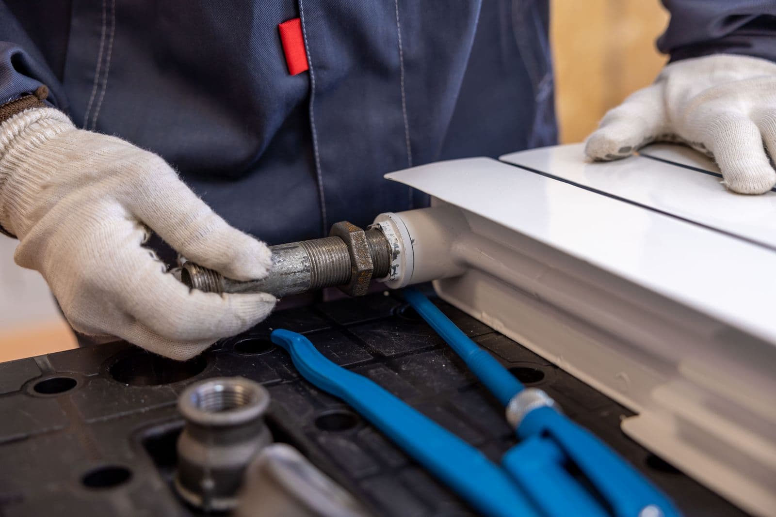 Gloved hands fitting a pipe connector to a white radiator, with blue pipe wrenches on the workbench.
